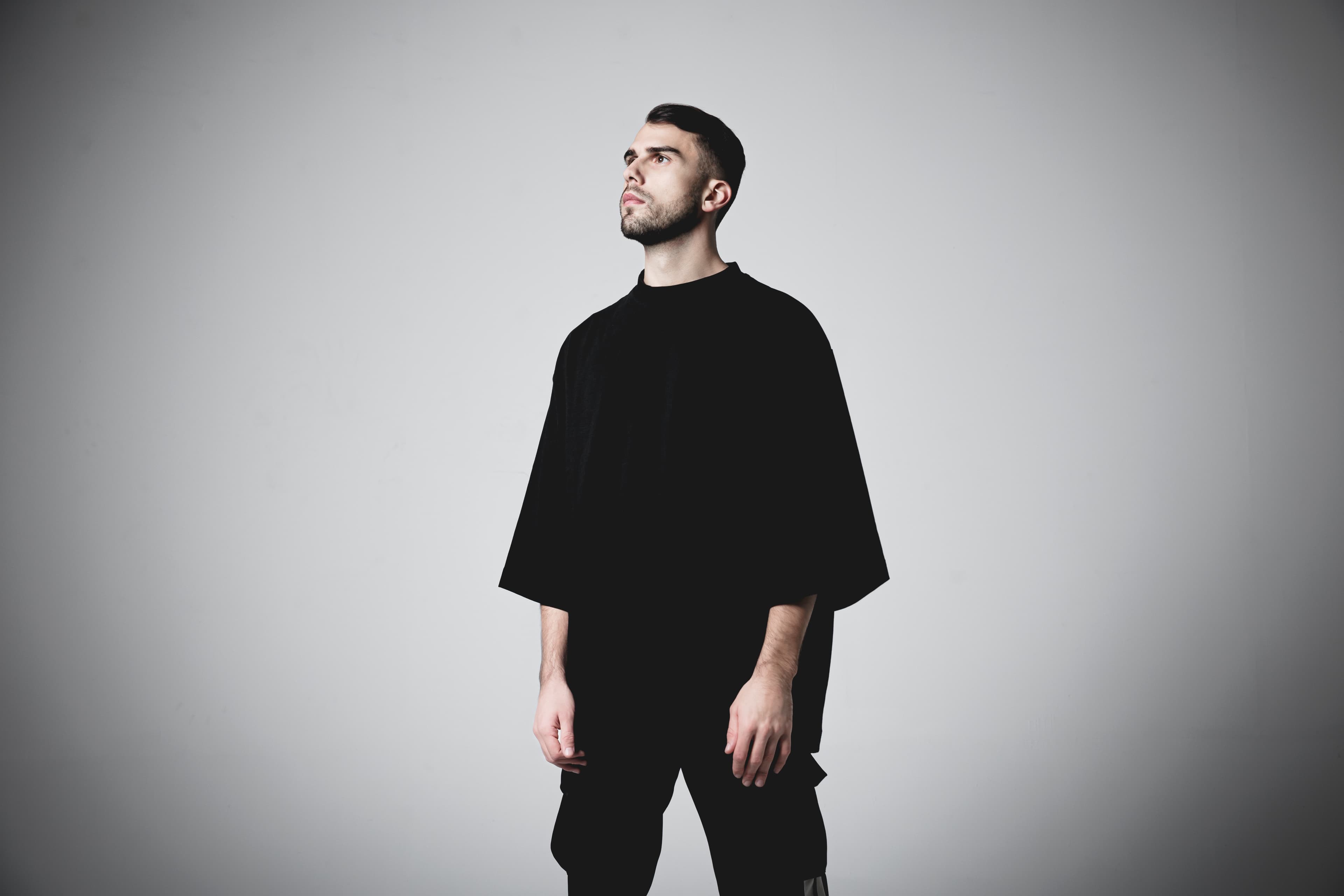 Bearded man in an oversized black t-shirt looking upwards against a plain grey background.